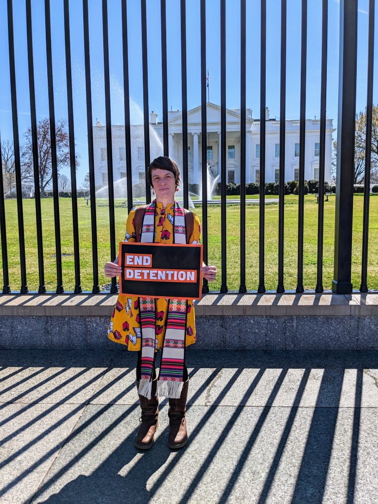 A woman with a clergy stole holding a sign that reads "End Detention" in front of the White House.